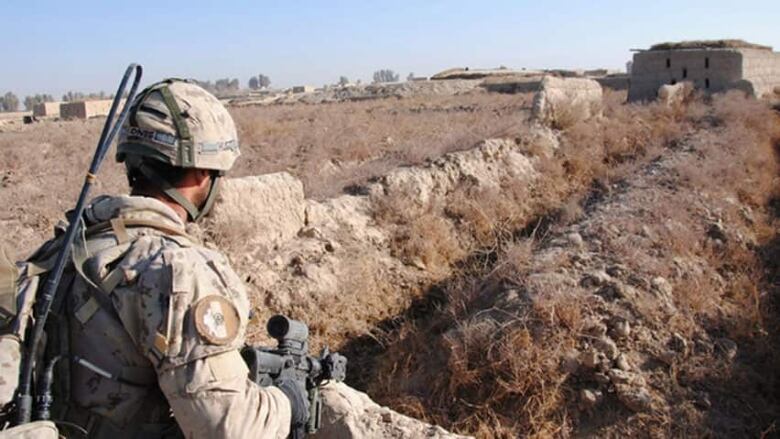 A solider, in desert camoflage and holding an assault rifle, looks at a building in the middle distance. 