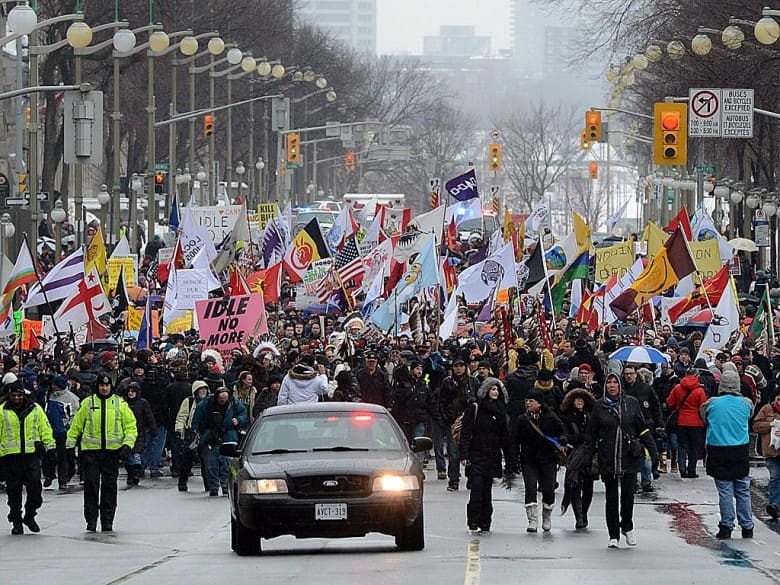 A large group of people, many carrying flags and banners, fill a street as they march behind a car with flashing lights.