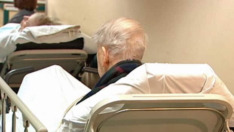 A man with tufts of white hair sits in a hospital bed, his back to the camera