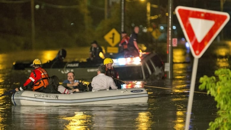 Emergency officials and stranded civilians take a life raft down a flooded city street, passing a YIELD sign on the way. It is dark, approaching evening