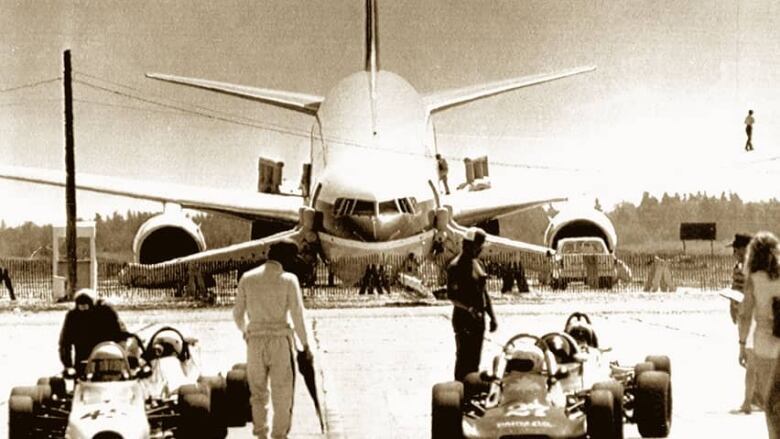 The front of plane that crashed on an airstrip. People next to race cars stand in front of it.