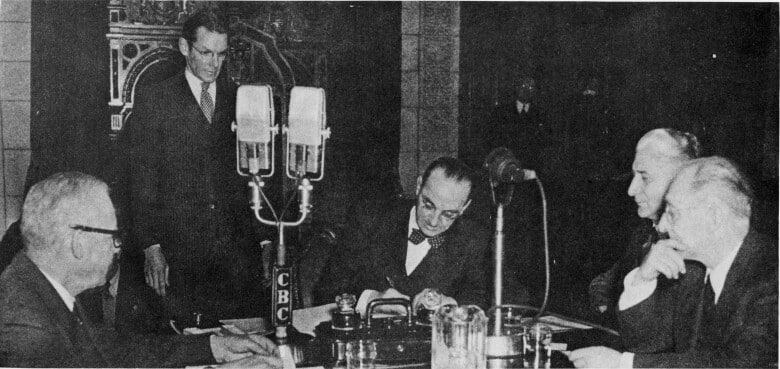 Black and white photo of group of men around a table, one is signing a document.