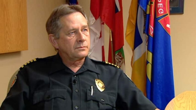 A man in a firefighter's uniform sits in a conference room with flags in the background behind him.