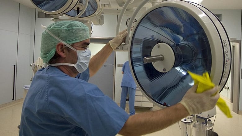 A man wearing hospital scrubs, a mask and gloves uses a yellow cloth to clean a piece of equipment in an operating room.