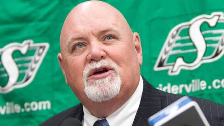 A man with a white goatee sits in front of a microphone with a Saskatchewan Roughriders logo behind him.