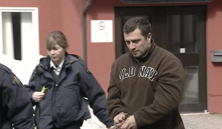 A man wearing a hoodie and restrained in handcuffs is led away from a courthouse by a female security officer in uniform.