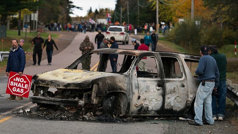 Protesters stand near a burned out vehicle lying on a street.