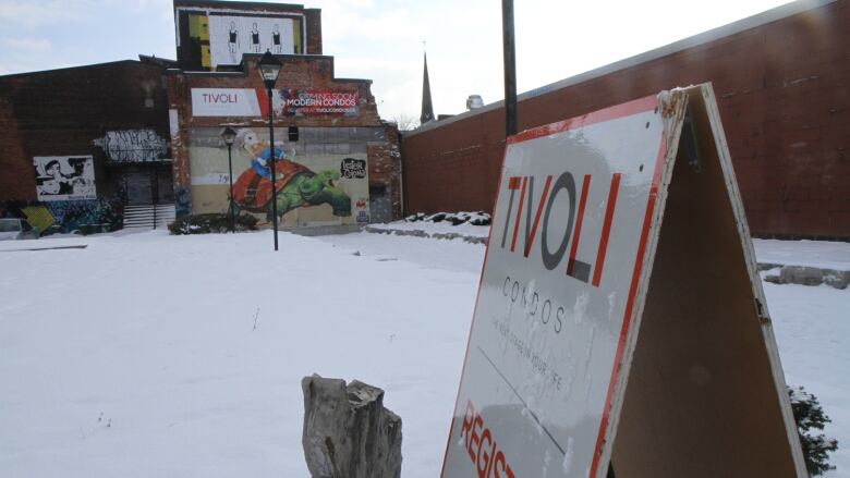 An empty lot covered in snow and a condo sign has an old brick building in the background.