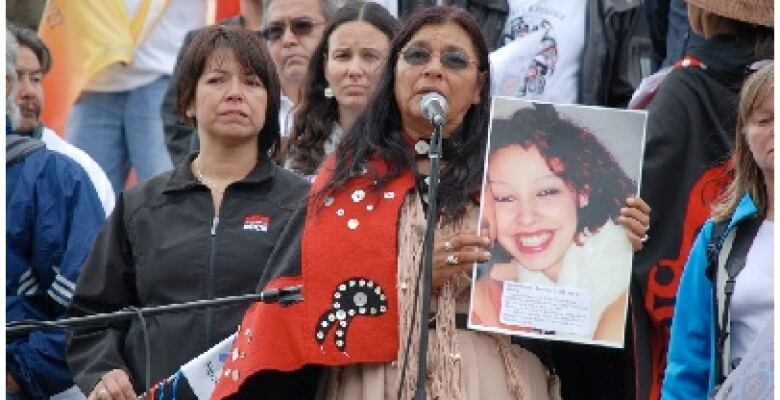 A woman in a crowd speaks at a microphone and holds a photograph of a young woman.