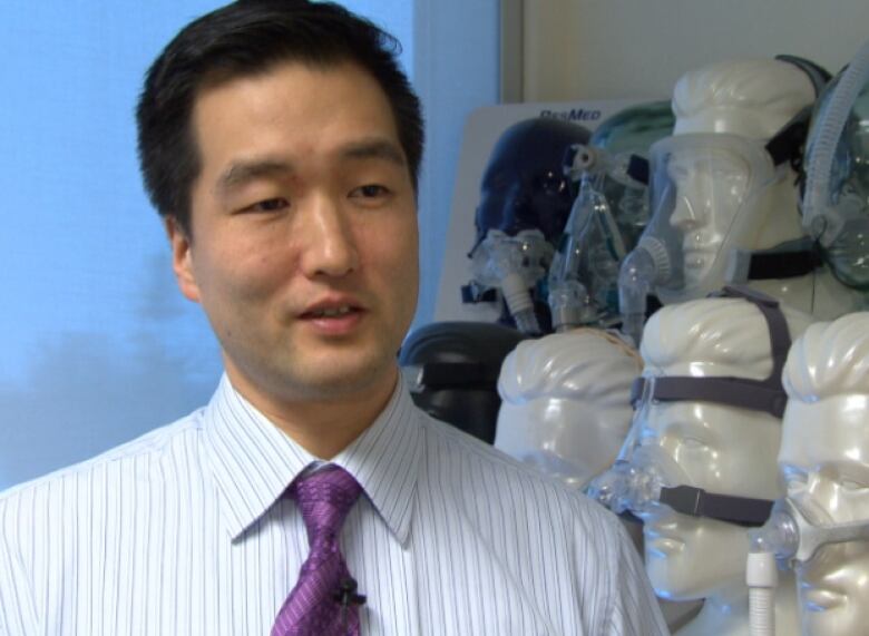 An East Asian man with short black hair, wearing a white shirt with thin blue stripes and a purple tie stands beside a row of mannequin heads with various breathing devices on their faces.