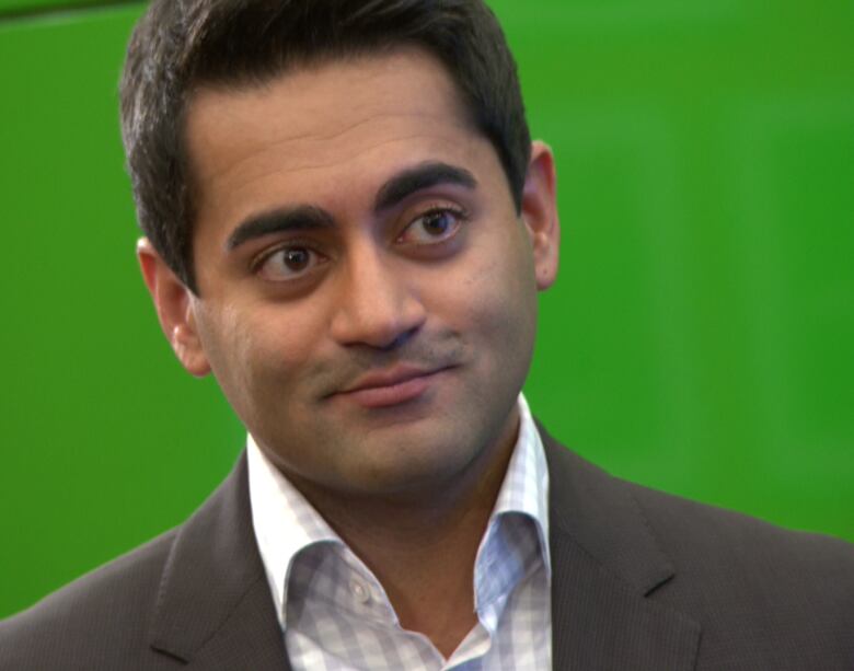 A man wearing a white checked shirt and grey blazer stands in front of a green screen.