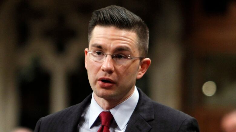 A man with glasses, wearing a dark suit and a red tie, stands to speak.