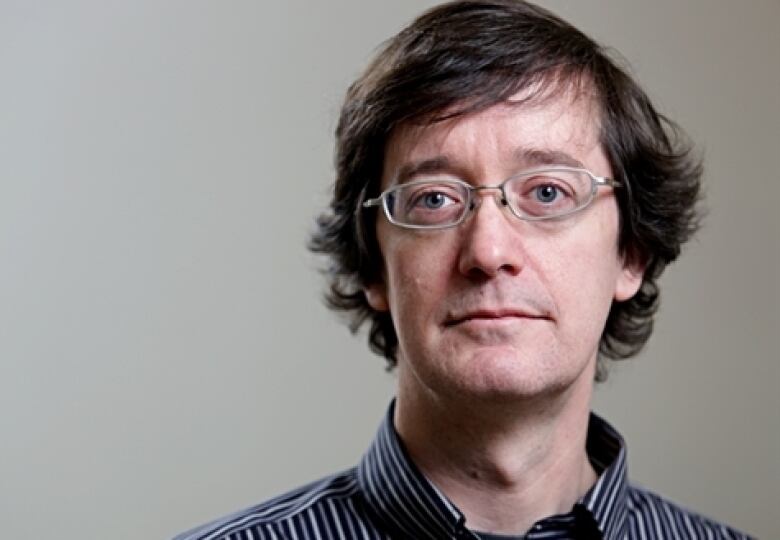 A man wearing glasses and a collared shirt poses for a photo in front of a grey background.