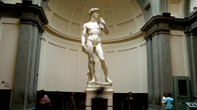 A marble statue of a nude man is seen in a museum as people walk by.