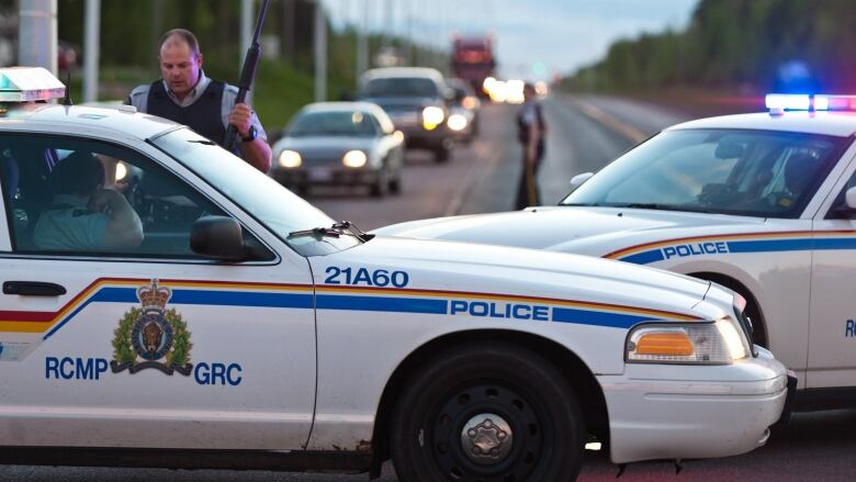 Two RCMP cruisers parked across a road with one officer standing outside holding a shotgun.