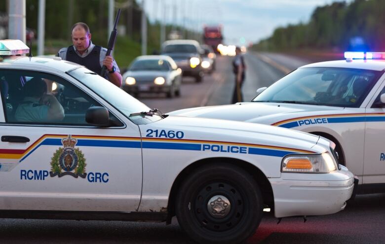 Two RCMP cruisers parked across a road with one officer standing outside holding a shotgun.