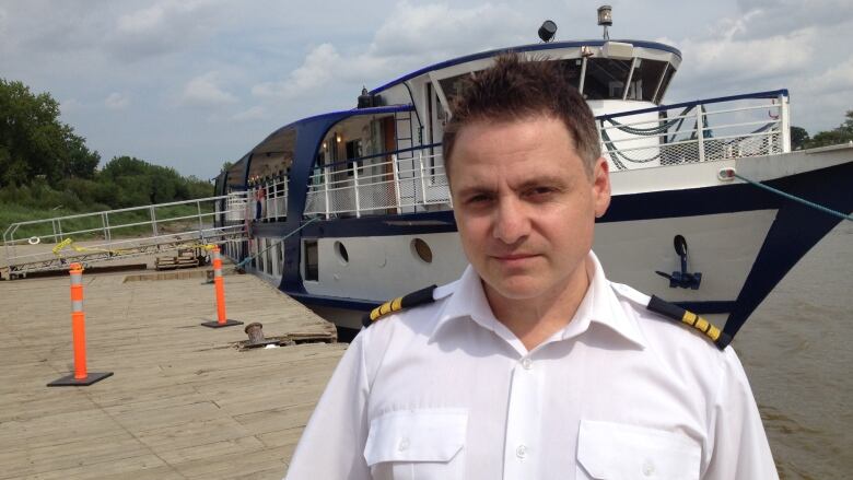 A man in a white uniform is seen standing on a dock with a small cruise ship behind him.