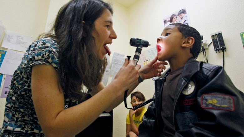 A health-care professional uses a lighted instrument to look in the mouth of a child on an examining table.