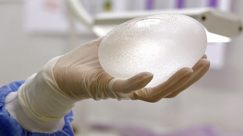 A gloved hand holds a silicone breast implant  a translucent, oblong structure about the same size as the hand.