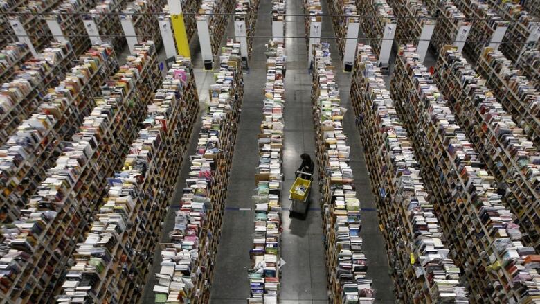 A person stands beside a cart on a warehouse floor among rows and stacks of books.