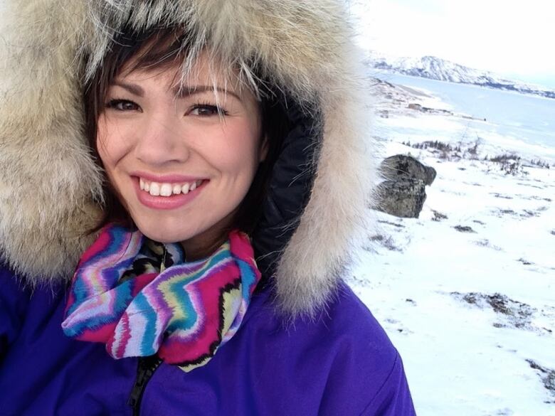 A smiling woman wearing a purple parka with a fur-lined hood stands outside, with a snowy expanse, with a body of water and hills behind her.