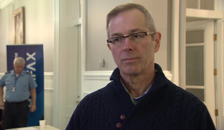 A white man with glasses stands in a hallway