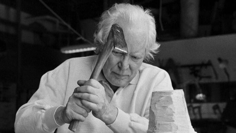 A black and white photo of a man with grey hair carrying a hammer as he carves a sculpture. 