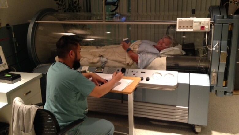 A woman lies in an enclosed bed in a hospital setting with a health care working sitting next to the machine.