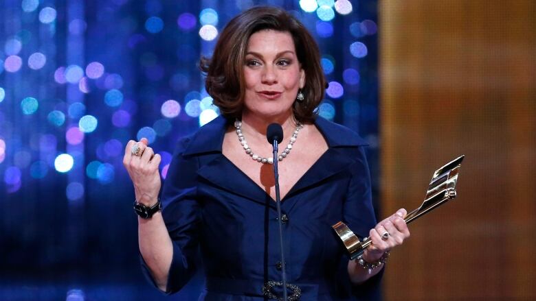 An elegantly dressed woman speaks into a microphone with an award statuette held in one hand. 