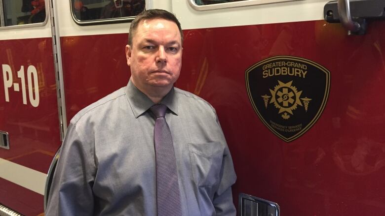 A straight-faced man in a shirt and tie stands next to a Sudbury fire truck