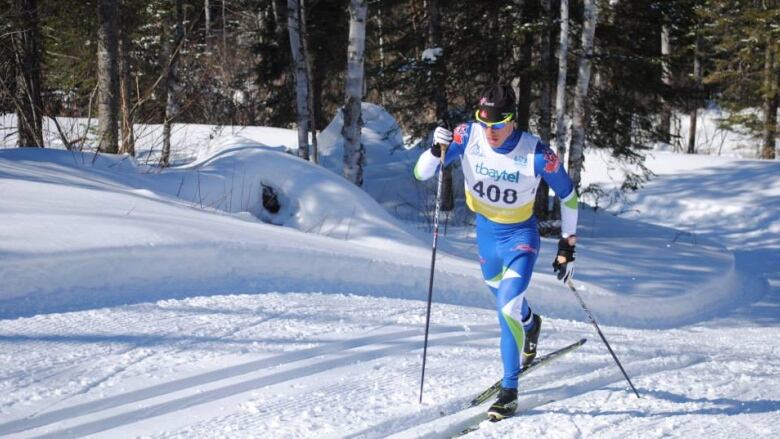 A man in a blue downhill suit and sunglasses cross-country skis up a hill in a wooded area.