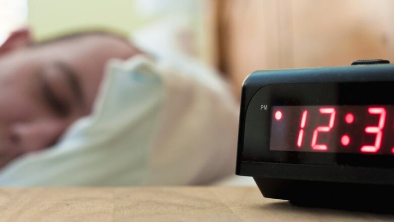 A man's head is seen resting on a pillow while a bedside alarm clock reads 12:30 p.m.