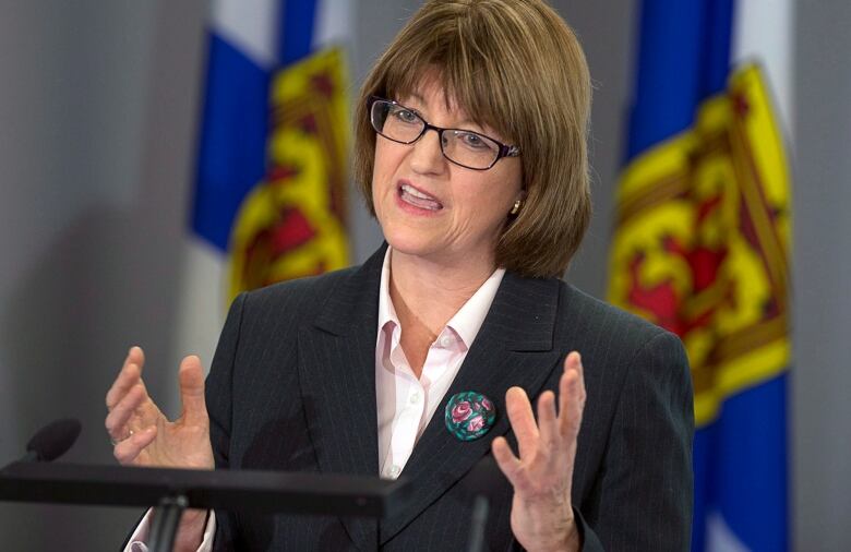 A woman with brown hair and glasses speaks at a podium, holding her hands up to explain a point. 