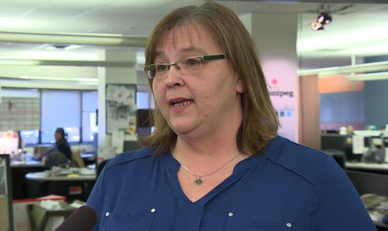 A woman in a blue shirt with glasses speaks into a microphone while standing in a newsroom.