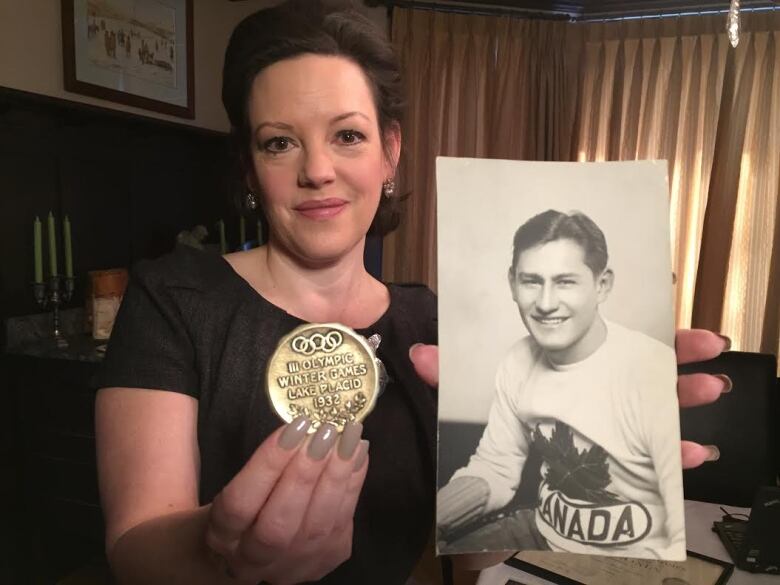 A woman is pictured holding up a black-and-white picture of a man in her left hand and a medal in her right hand, while looking forward to the camera.