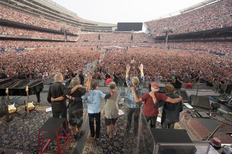 Seven members of the Grateful Dead stand on stage with their backs to the camera looking out at a huge crowd of people in a stadium. 