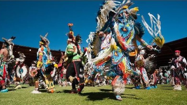 A number of people in Indigenous regalia dance on a grassy area.