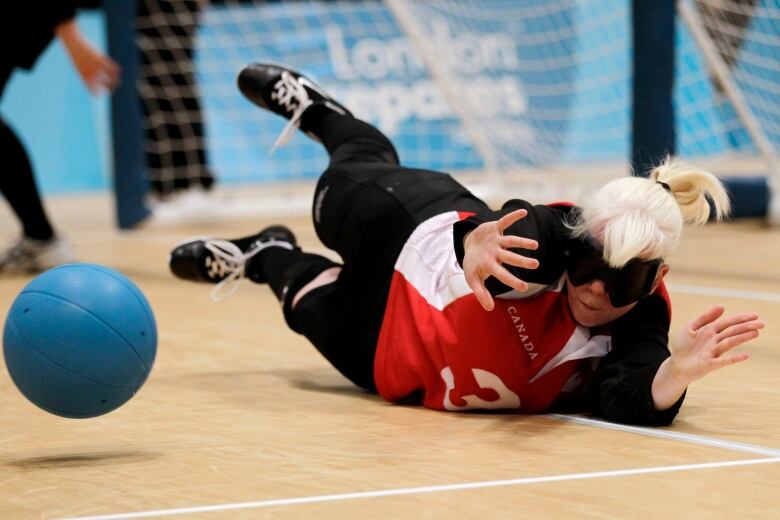 A woman on a gymnasium floor dives and puts out her hand to stop a blue ball. 