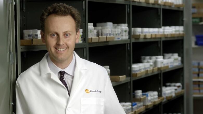 A man in a white lab coat leans against a shelf holding various pill bottles and smiles.