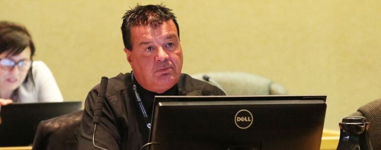 A man with black hair, wearing a black shirt sits at a table.