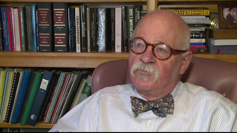 A man with glasses and a moustache sits in front of a bookshelf.