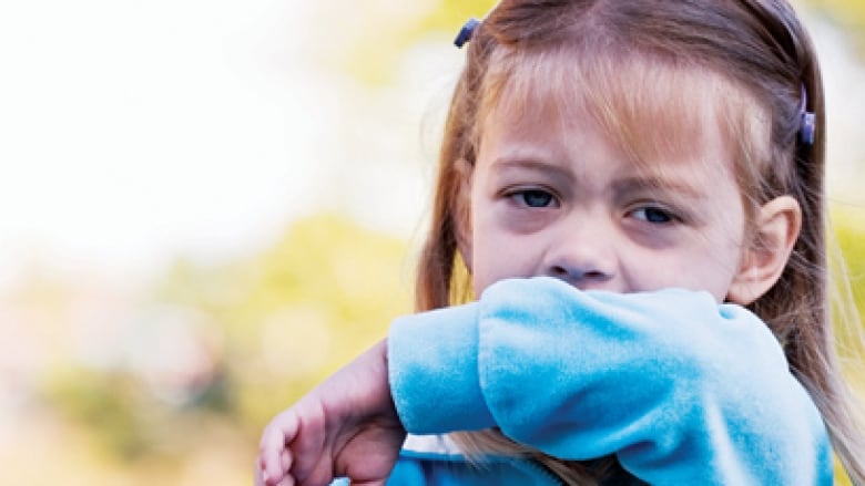 A young girl covers her mouth with her arm as she coughs.