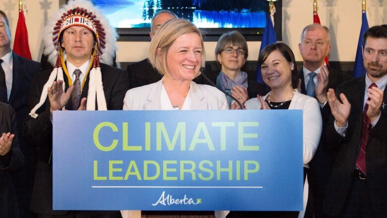 Rachel Notley, smiling, stands behind a podium that says 'Climate Leadership,' with a number of industry, environmental and Indigenous leaders clapping.