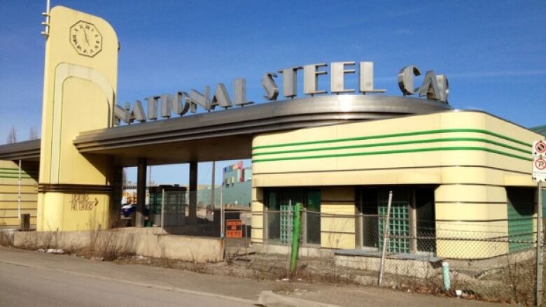 An art deco building and clock that says National Steel car is surrounded by chain link fence.