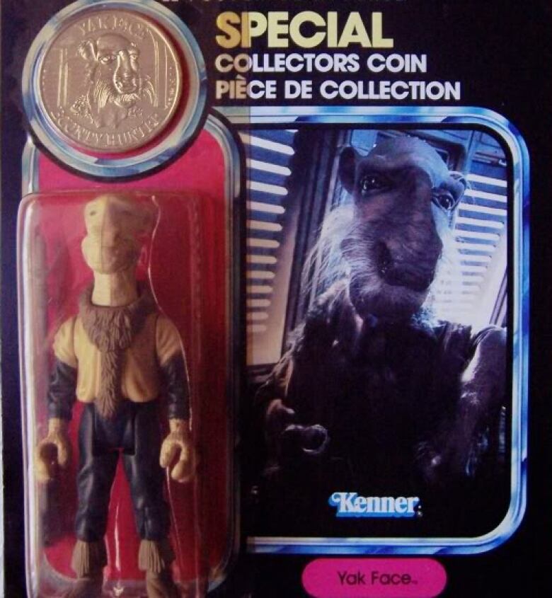 An alien figurine stands in its clear plastic packaging with a coloured backdrop. There is a coin above his head. The packaging says 