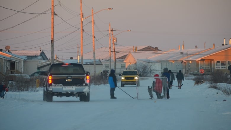 A truck veers around kids playing road hockey on a snow-covered street with houses basked in the sun at dusk. 