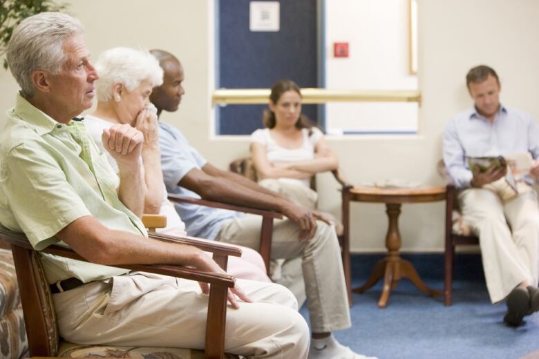 Five adults, including two seniors in the foreground, sit waiting in a doctor's office waiting room.