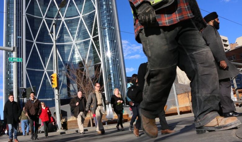 People wearing work boots and business attire walk across a busy downtown crosswalk. In the background is a large office tower. 