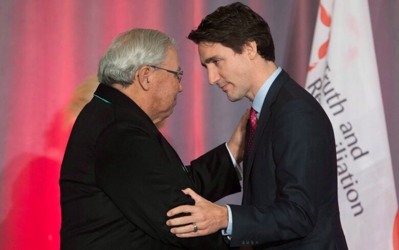 Two men shake hands in front of a flag that reads 'Truth and Reconciliation.'