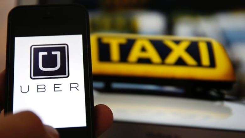 A person holds up a phone with the word 'Uber' on it next to a taxi sign.